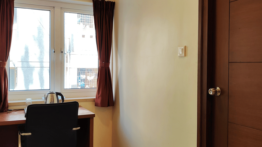 A well-lit room featuring a small wooden desk and chair positioned near a window, with a door leading into the main area | A & M Rooms and Residences