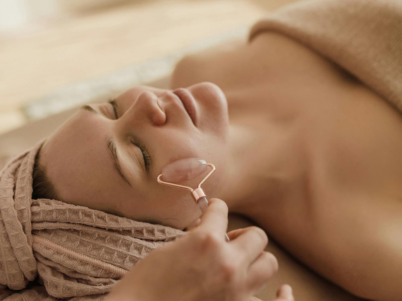 A woman with a towel wrapped around her head is lying down with her eyes closed as a hand uses a pink crystal facial roller on her cheek.