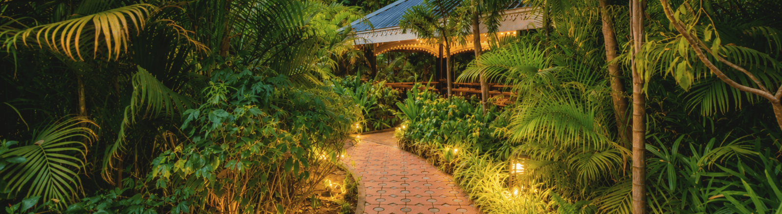 Garden pathway lit at dusk winding through dense tropical foliage towards a gazebo at MAYFAIR Oasis Resort & Convention, Jharsuguda.