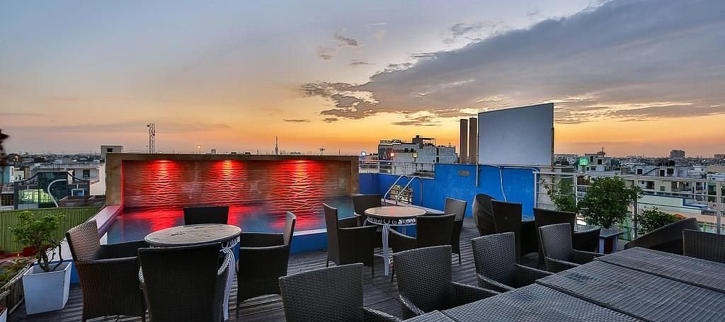 A rooftop terrace during sunset with wicker furniture, a lit pool and a view of the city skyline - TGI Apple Inn, Jaipur 