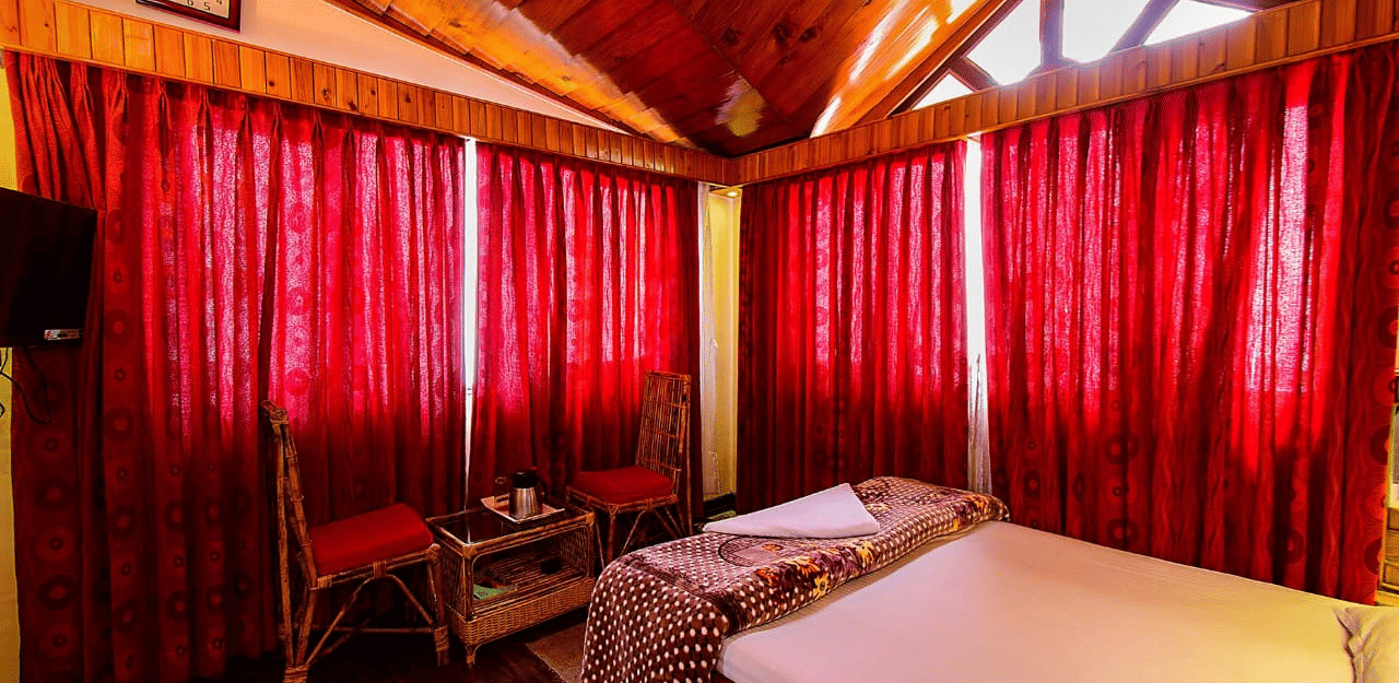 Close-up of a double bed with a brown patterned throw, 2 wicker chairs, red curtains, and a wooden vaulted ceiling at Hotel Dolphin Darjeeling.