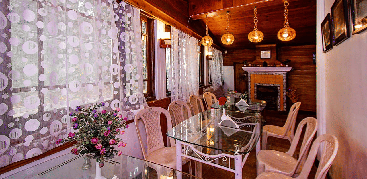 Dining area with a wooden ceiling, hanging lights, a glass table, and floral patterned white lace curtains at Hotel Dolphin Darjeeling.