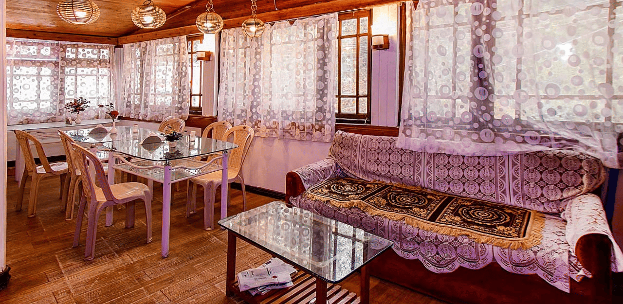 Restaurant/dining room with lace curtains, wooden walls and ceiling, glass tables, and light-coloured chairs at Hotel Dolphin Darjeeling.