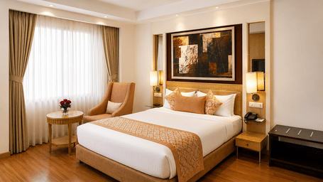 Hotel room with a bed dressed in white and gold linen, a beige armchair, a wall-mounted artwork and long sheer curtains by the window nearby.