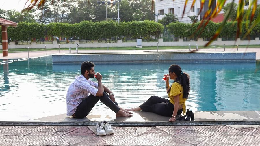 A man and a woman sit on the edge of a pool facing each other, talking and smiling. Trees and buildings are visible in the background at Jenneys Residency
