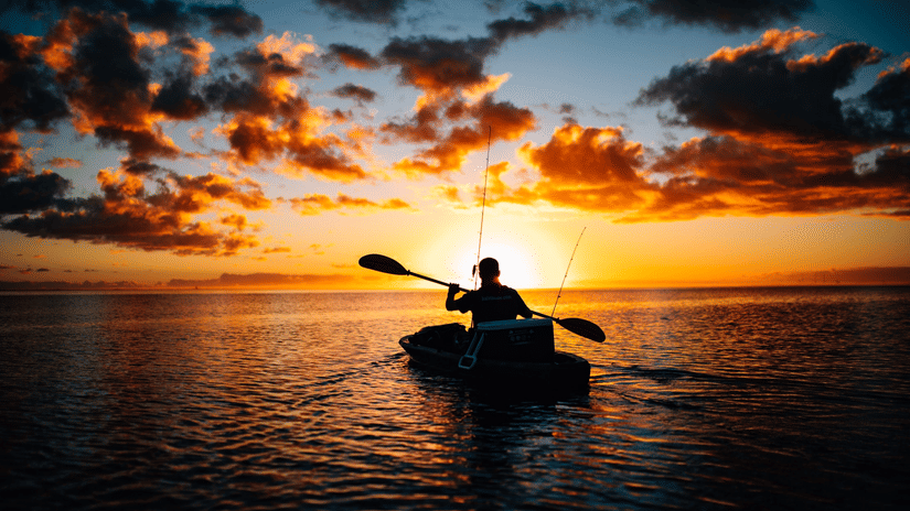 a person kayaking on a water body with the sun setting in the background making the sky turn into hues