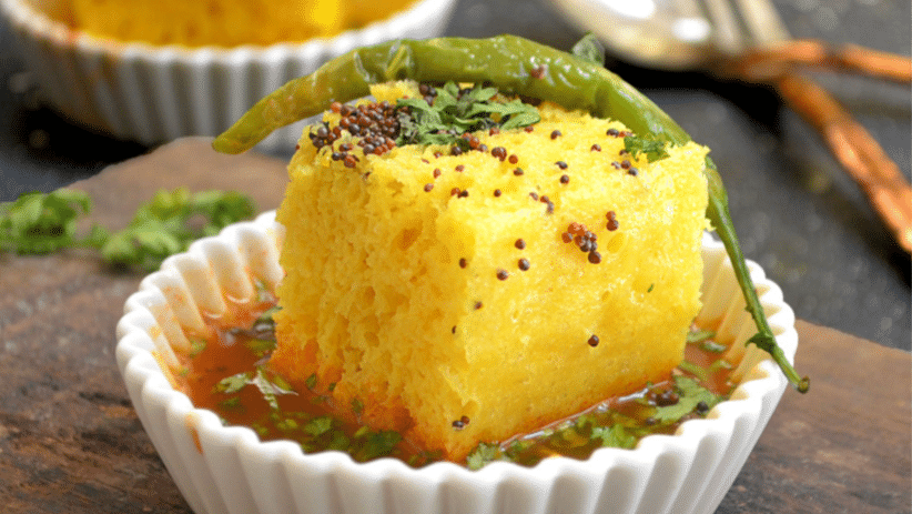 Dhokla served in a small bowl, garnished with fresh coriander and fried green chili, resting on a bed of vibrant chutney.
