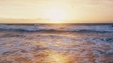 A serene ocean scene at sunset with soft waves breaking on the shore and a glowing sky in the background.