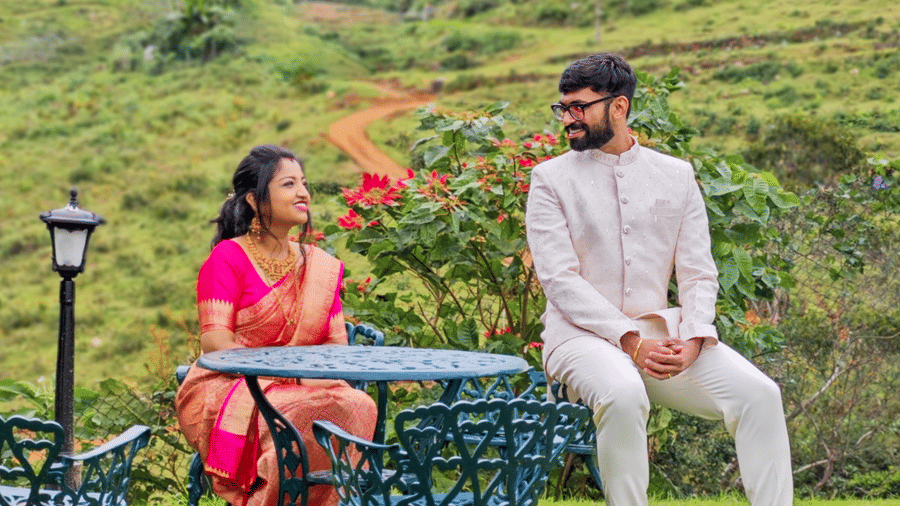 A couple is seated at a small metal table on a grassy lawn with a scenic hilly background; the woman wears a pink sari, and the man wears a white suit.