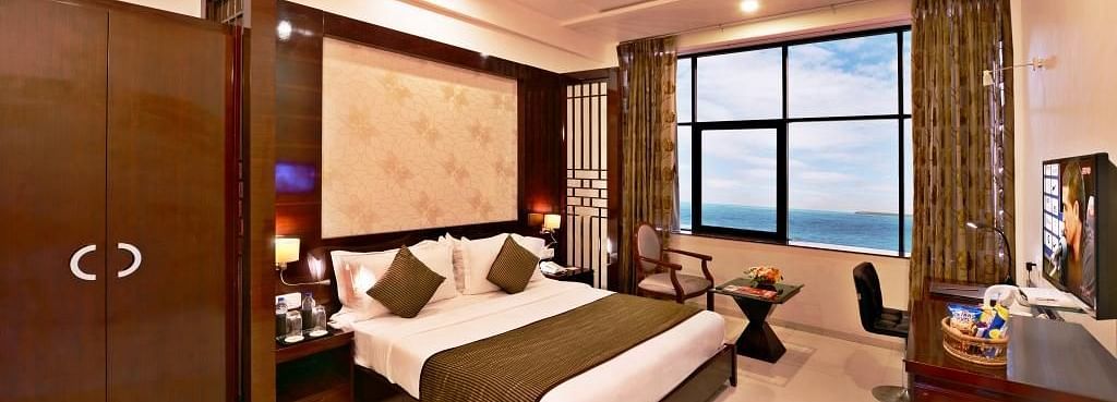 A cosy hotel room includes a king size bed with patterned bedding, a seating area near a window with a view, and warm lighting.
