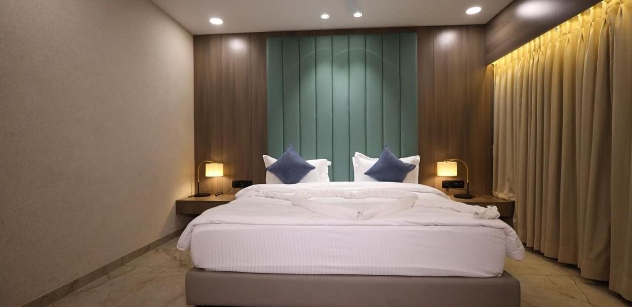 The image shows a bedroom with a bed, pillows, bedside tables with lamps, and curtains - Lords Eco Inn Navsari.