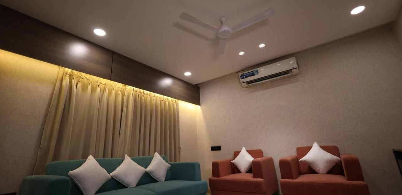 The image shows a living room with a sofa, two armchairs, curtains, and ceiling lights - Lords Eco Inn Navsari.