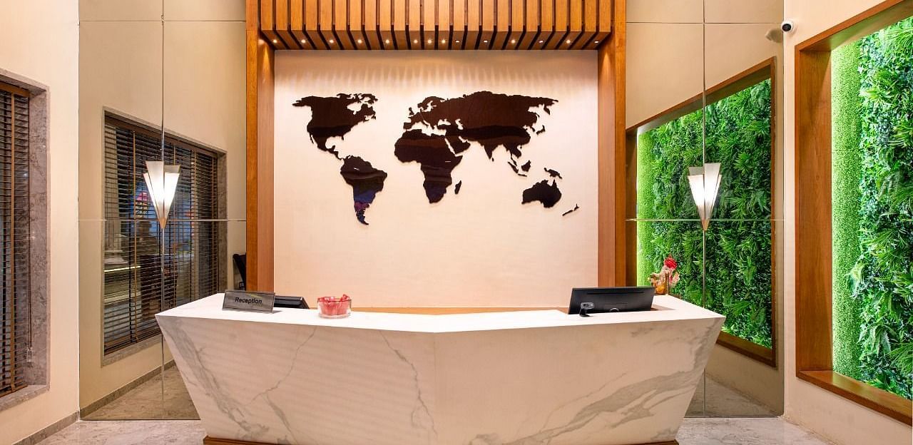 A contemporary reception area with a sleek white and gold front desk, a large dark world map on the wall, vertical wooden slats above, and illuminated green moss panels on the side walls, creating a modern and welcoming atmosphere - Lords Inn Motikhavdi, J