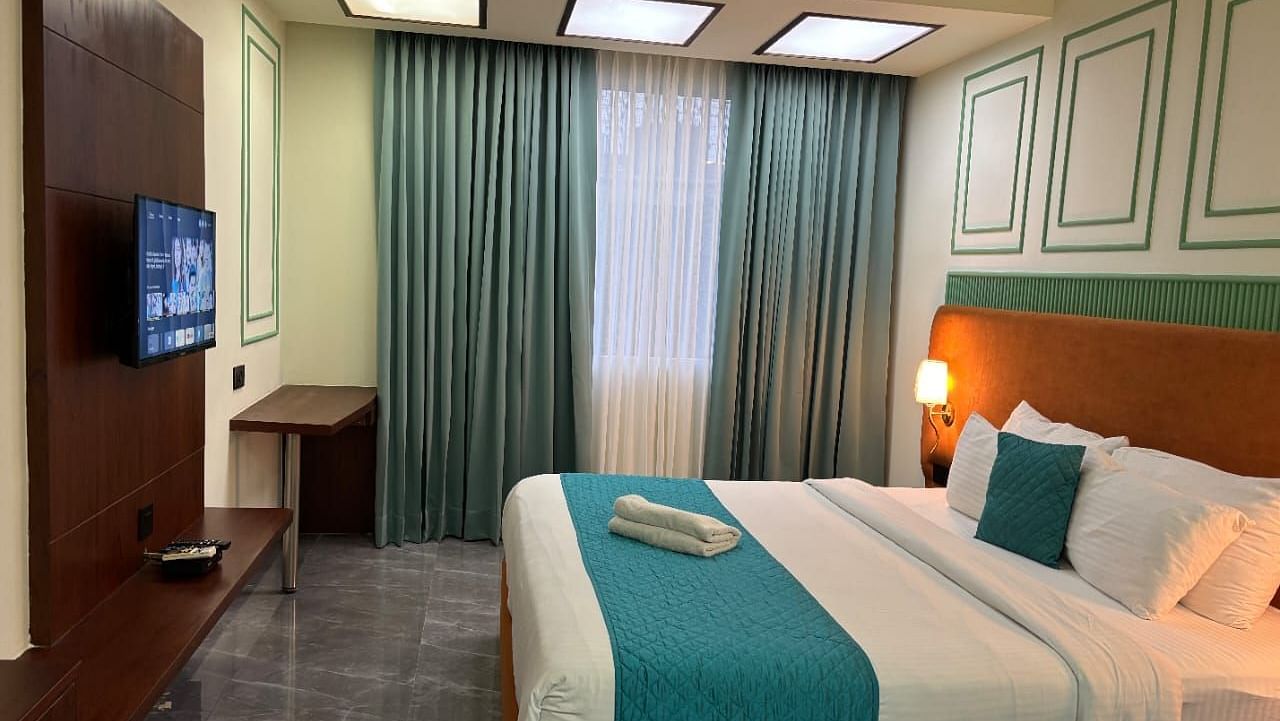 A comfortable room showing a double bed with blue accents, a television, and curtains - Asian Suites IFFCO Metro, Gurgaon