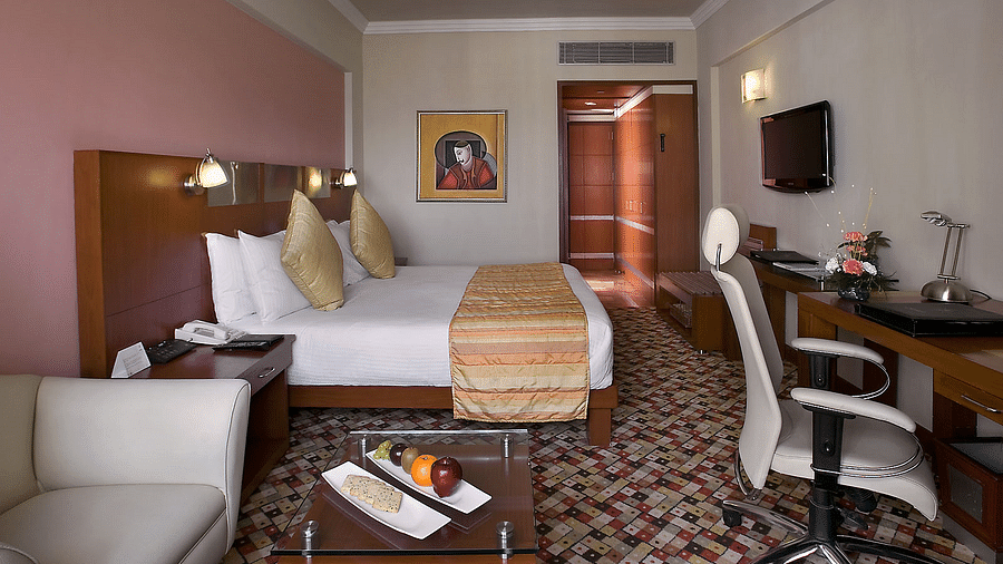 An image of a Deluxe Room with double size bed, a sofa and centre table, flat TV screen on the wall, and a work desk with chairs -The Suryaa New Delhi