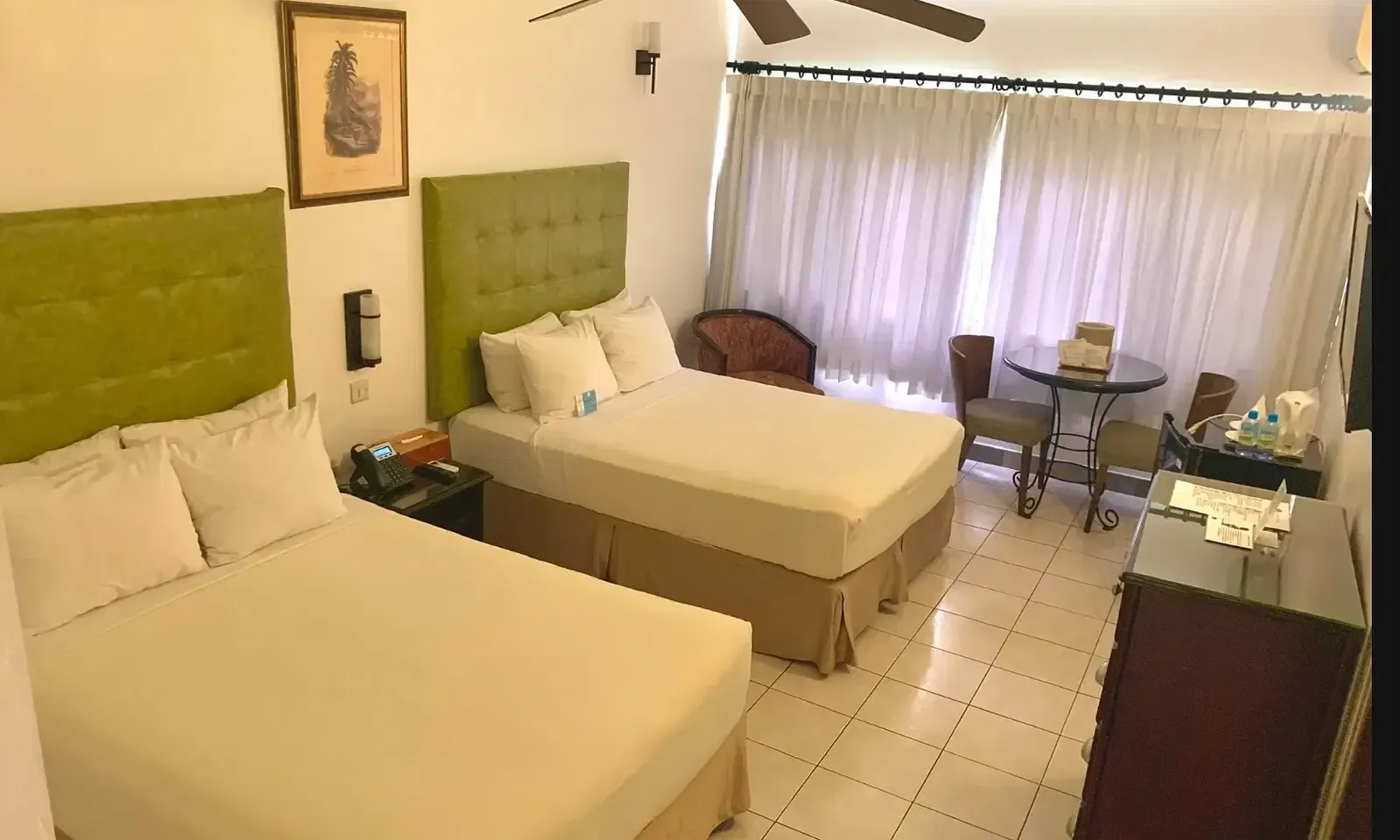 A hotel room showcases two beds with green headboards, a dresser, and a television, with a ceiling fan visible | Executive Double Room | Altamont Court, Jamaica