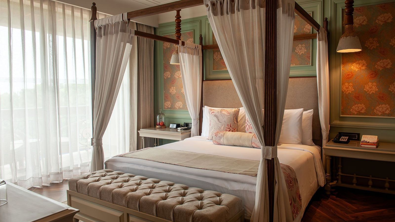 A bright and inviting suite at The Evren, Vagator, with a four-poster bed, white flowing curtains, and a plush tufted bench. The room features intricate wallpaper details and a stylish wooden floor.