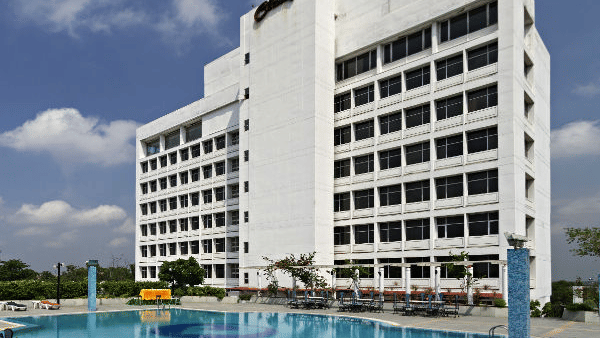 A large, modern white hotel building is shown behind a spacious blue swimming pool with lounge chairs on the patio - Clarks Avadh, Lucknow