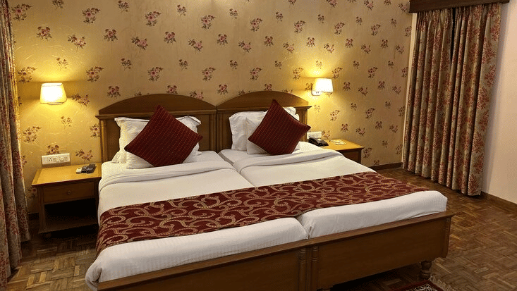 A bedroom with win beds with maroon pillows against a floral patterned wallpaper background at Amantra Shilpi Resort, Udaipur.