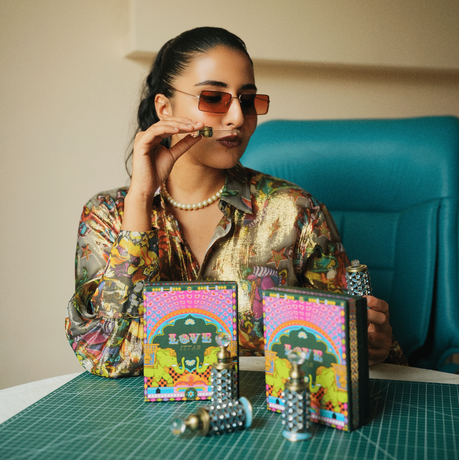 A stylish woman in sunglasses and a shiny shirt holds a perfume bottle, seated at a table with colourful perfume boxes labeled 'LOVE' and decorative vials - Kastoor x Sawai Hospitality