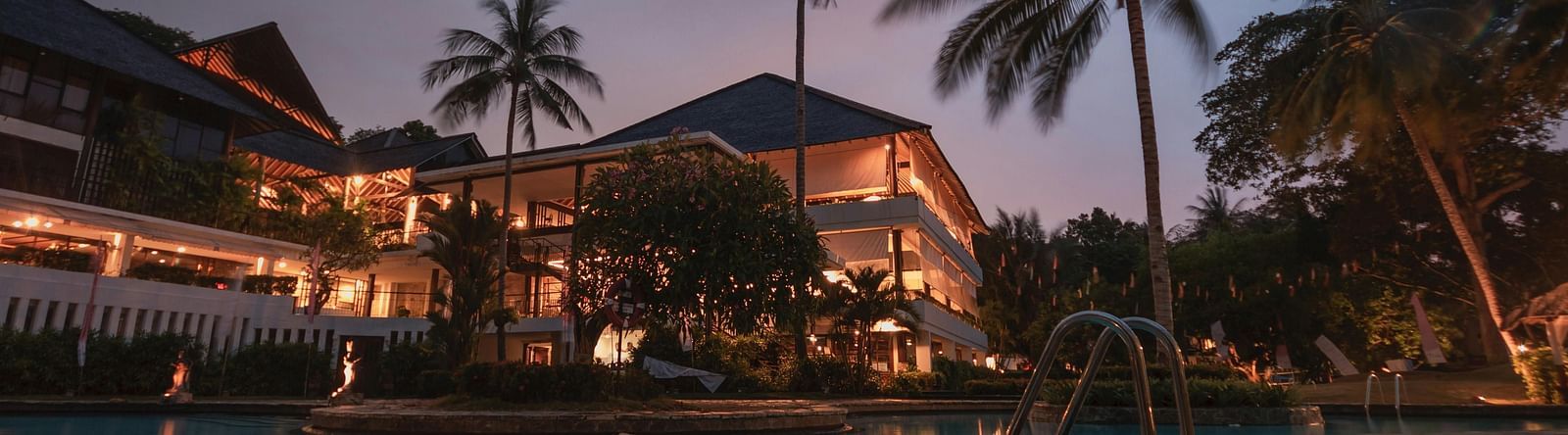  A hotel facade against the evening sky, surrounded by coconut trees near the pool, with the building lights reflecting on the water