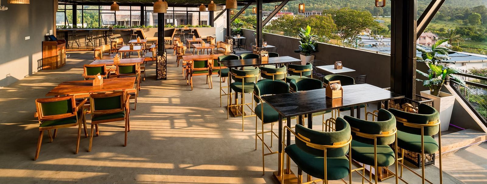 A spacious rooftop restaurant with elegant wooden furnishings, green velvet chairs, and panoramic views of the hills, bathed in warm natural light at TGI Livancia Hillside, JC Nagar, Mysore.
