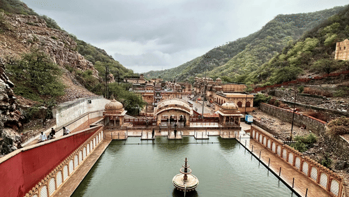 A fenced pond at the Galta Ji Temple in Jaipur surrounded by the Aravalli Hills under a cloudy sky.