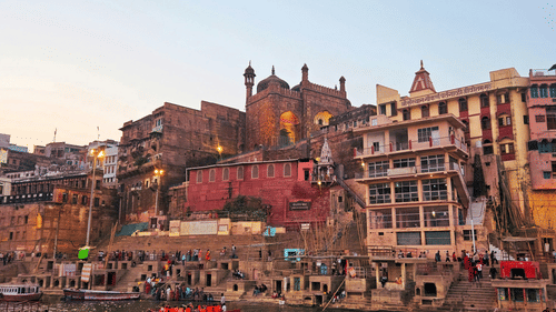 A long shot of a boat with passengers on the Ganges River, moving past the Varanasi ghats with buildings and people visible along the riverbank with clear sky in the background.