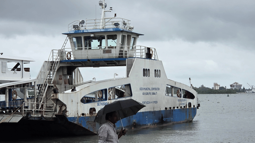 Person walking along a wet pier holding umbrella near a ferry under cloudy monsoon skies at harbour in coastal town