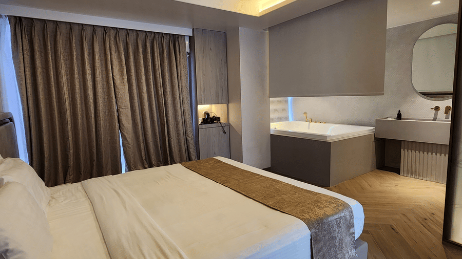 Modern Smart Room with Double Seater Bathtub room at Dragonfly Hotel – The Art Hotel with large bed, white linens, brown runner, cushioned headboard, bench at foot of bed, floor?to?ceiling curtains, recessed tray ceiling lights, and vanity area with mirror