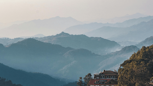 Layered mountain ranges recede into the mist under a soft blue sky, while a building with a red roof perches on the hillside, integrating human habitation into the tranquil landscape.