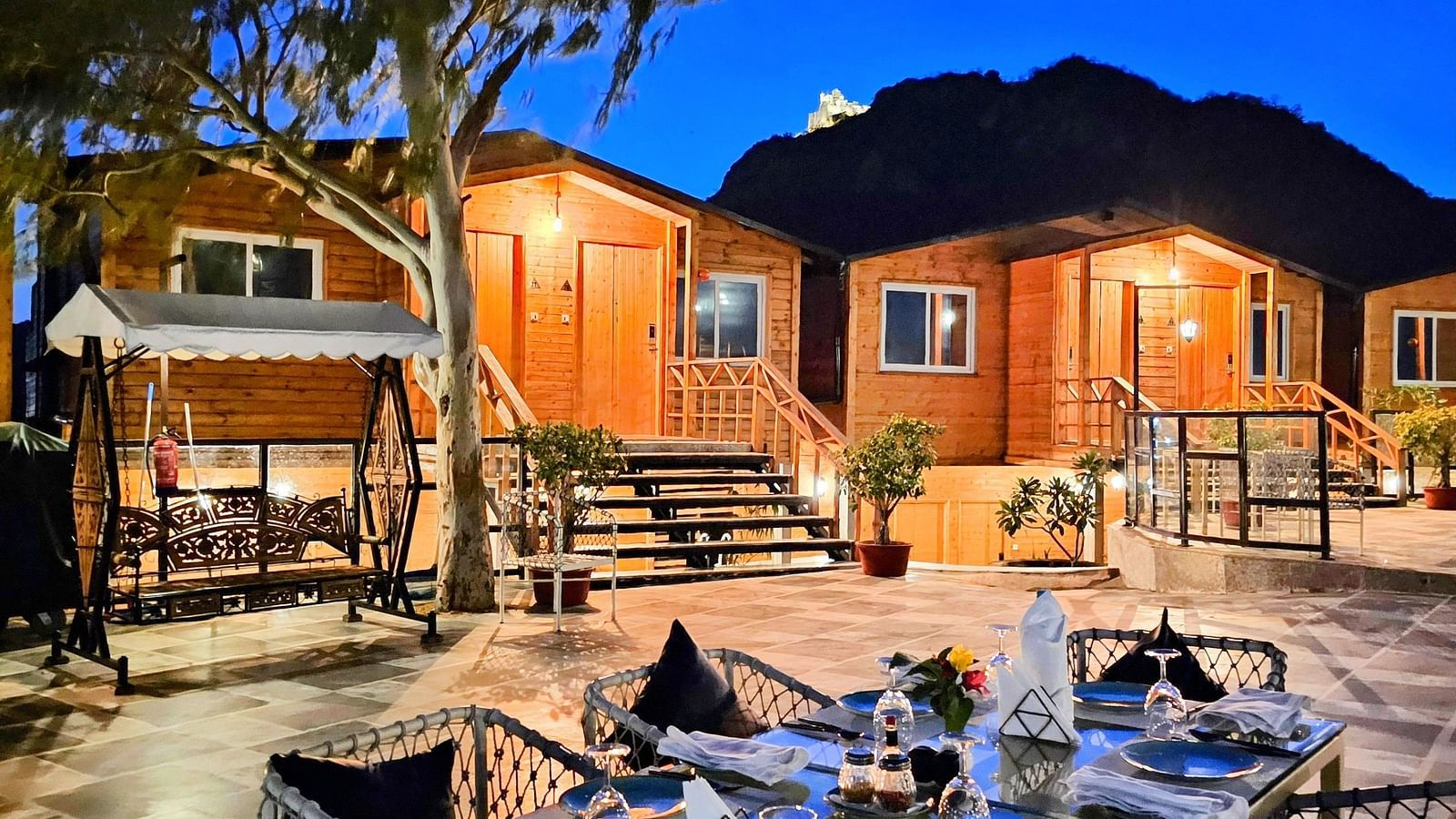 An outdoor dining table with wooden cabins in the background. - Bamboo Saa Resort & Spa, Udaipur.
