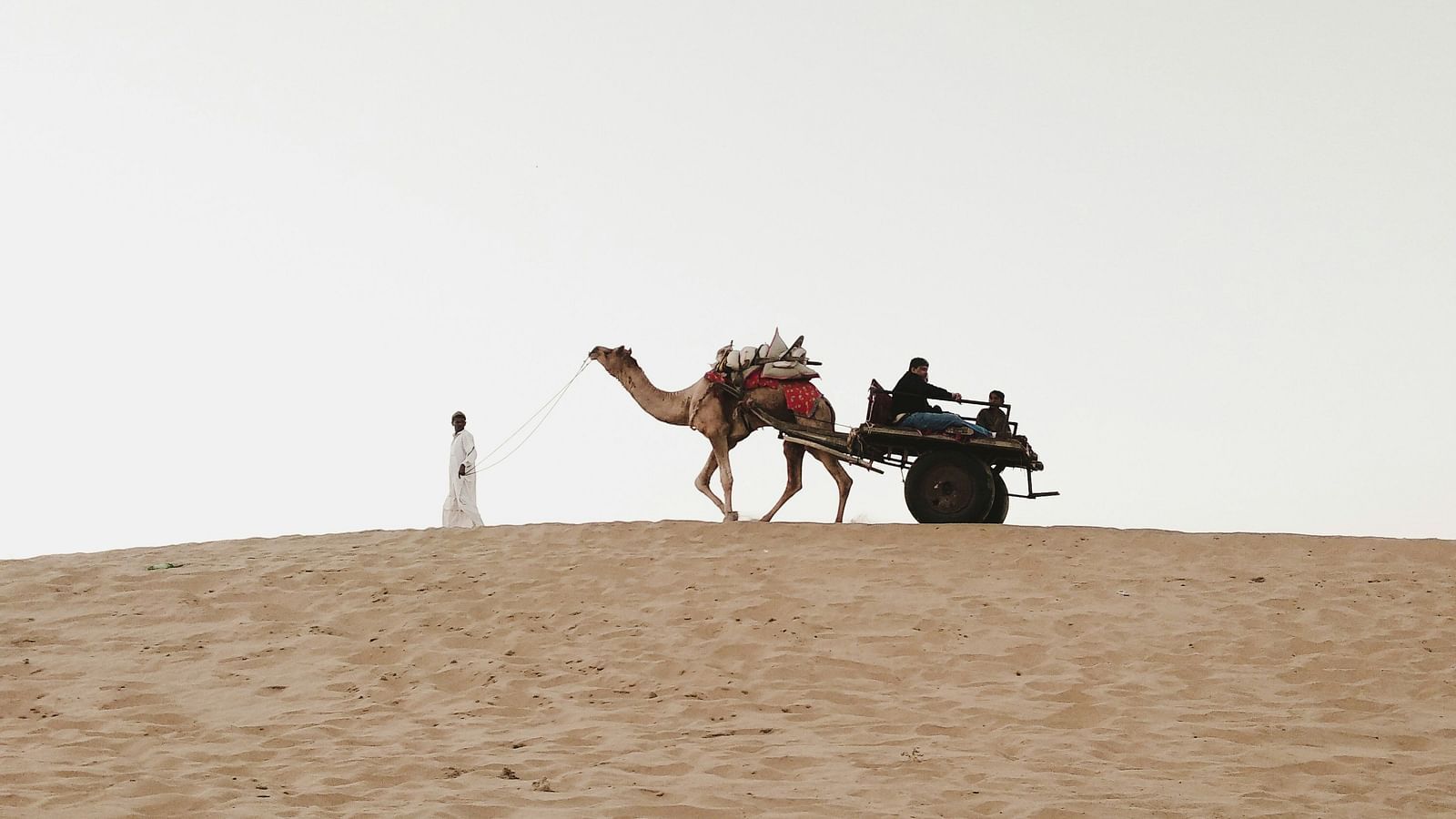 A person walking in Thar Desert and taking a camel with another passenger sitting behind.
