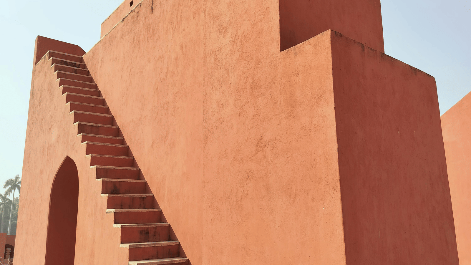 A large, stepped, terracotta-coloured masonry structure with a broad diagonal staircase, likely a massive sundial or other astronomical instrument from the Jantar Mantar observatory, is photographed against a bright, hazy sky.