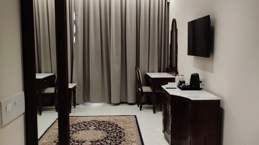 A room at Anant Mahal, Jaipur featuring a large mirror, a patterned rug on the floor, a large window covered by blinds, and a wall mounted television.