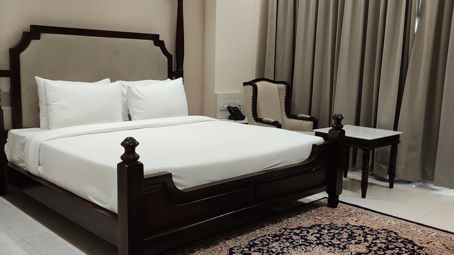 A bedroom at Anant Mahal, Jaipur featuring a king size bed, a patterned rug, floor to ceiling curtains behind a table, and a chair.