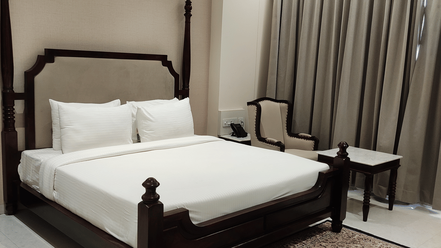 A bedroom at Anant Mahal, Jaipur featuring a king size bed, a patterned rug, floor to ceiling curtains behind a table, and a chair.