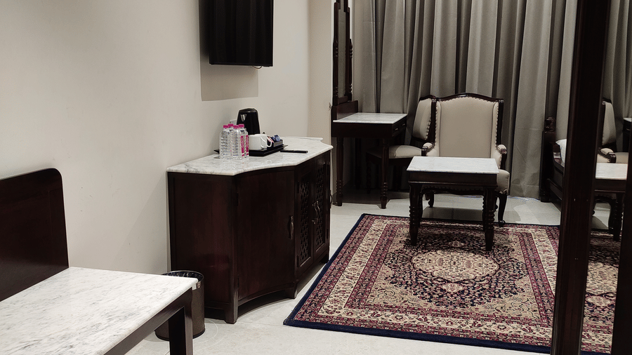 A room at Anant Mahal, Jaipur with a wooden cabinet, a wall mounted television, a patterned rug on the floor, a table and a chair.