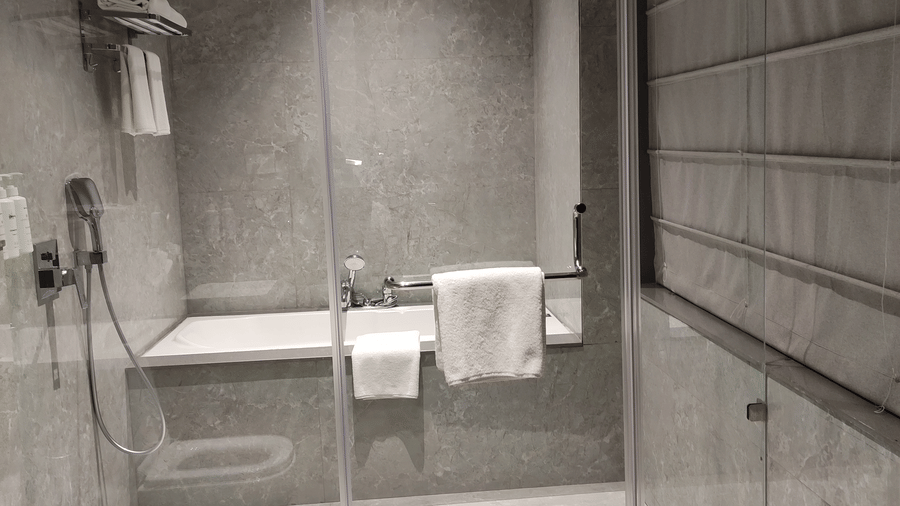 A grey tiled bathroom at Anant Mahal, Jaipur featuring a glass shower door, a white toilet, and 2 white towels hanging on a metal rail.