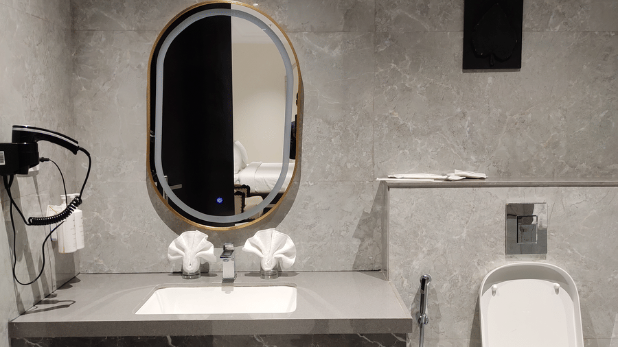 Bathroom interior at Anant Mahal, Jaipur with a stone vanity, an oval mirror, a wall mounted hair dryer, and a white toilet.