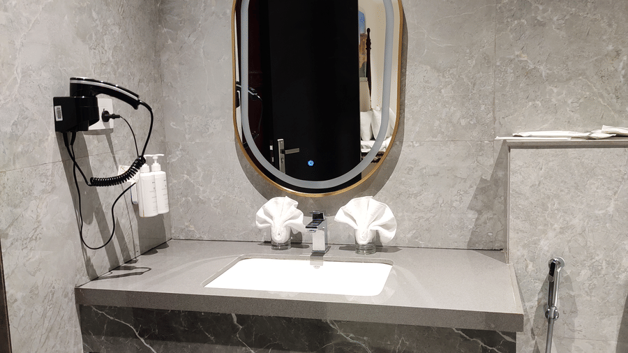 A bathroom at Anant Mahal, Jaipur featuring a stone countertop with a sink, open wooden shelving, a hair dryer, and an oval mirror.