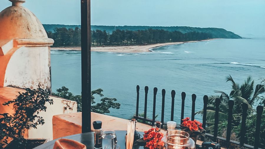 An outdoor dining table on a balcony at Fort Tiracol, Goa, set with food and drinks, overlooking a scenic view of a bay and distant land under a light sky.