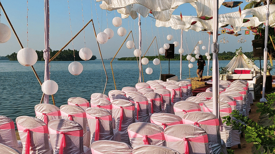 Rows of white chairs with pink covers arranged outdoors under hanging umbrellas at Paradise Lagoon Resort, Udupi.