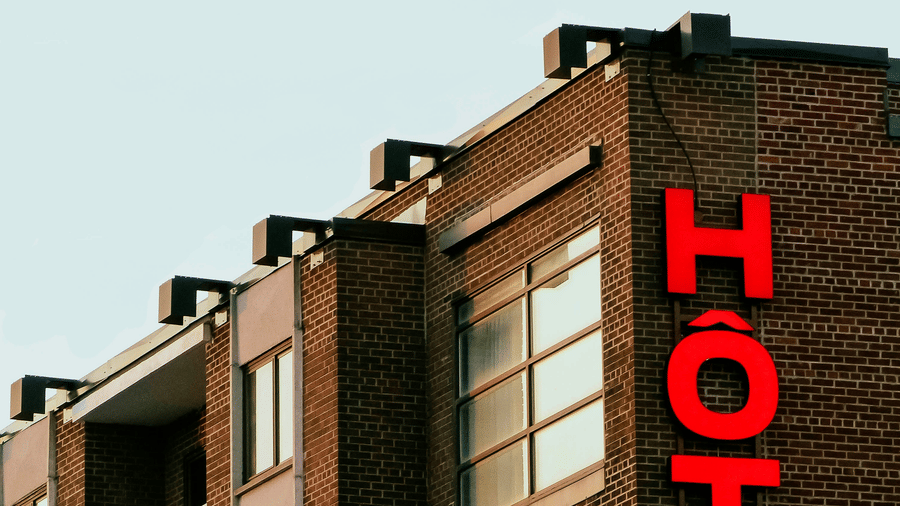 The exterior corner of a brick hotel building featuring a prominent vertical red neon sign.