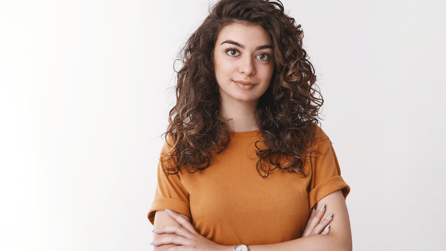 Woman with curly hair wearing an orange top, standing with arms crossed.