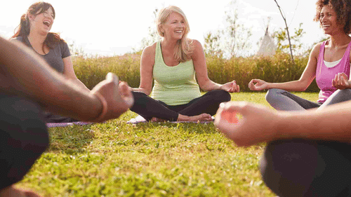 A diverse group of five women smiles and laughs while sitting in a circle on yoga mats outdoors, enjoying a sunny day at YO1 Longevity & Health Resorts, Catskills
