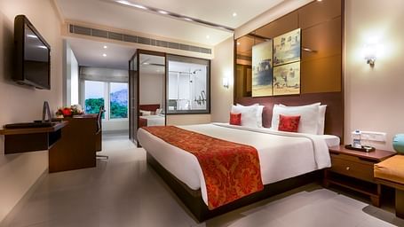 Hotel room with a king-sized bed, red bedding, and a sofa seating area. There's a TV on a stand, artwork on the wall, and a door leading to a bathroom.
