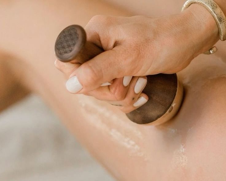 A hand holding a wooden massage tool and applying it to a person's leg, which has some oil or lotion on it.