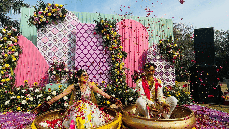 a bride and groom sitting on big bowls during haldi ritual with rose petals placed around them - heritage village resorts & spa, Manesar