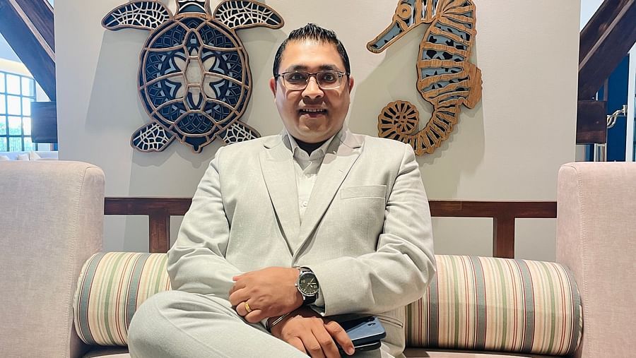 A person wearing a light-colored suit sits on a cushioned bench in front of a wall decorated with nautical-themed items.